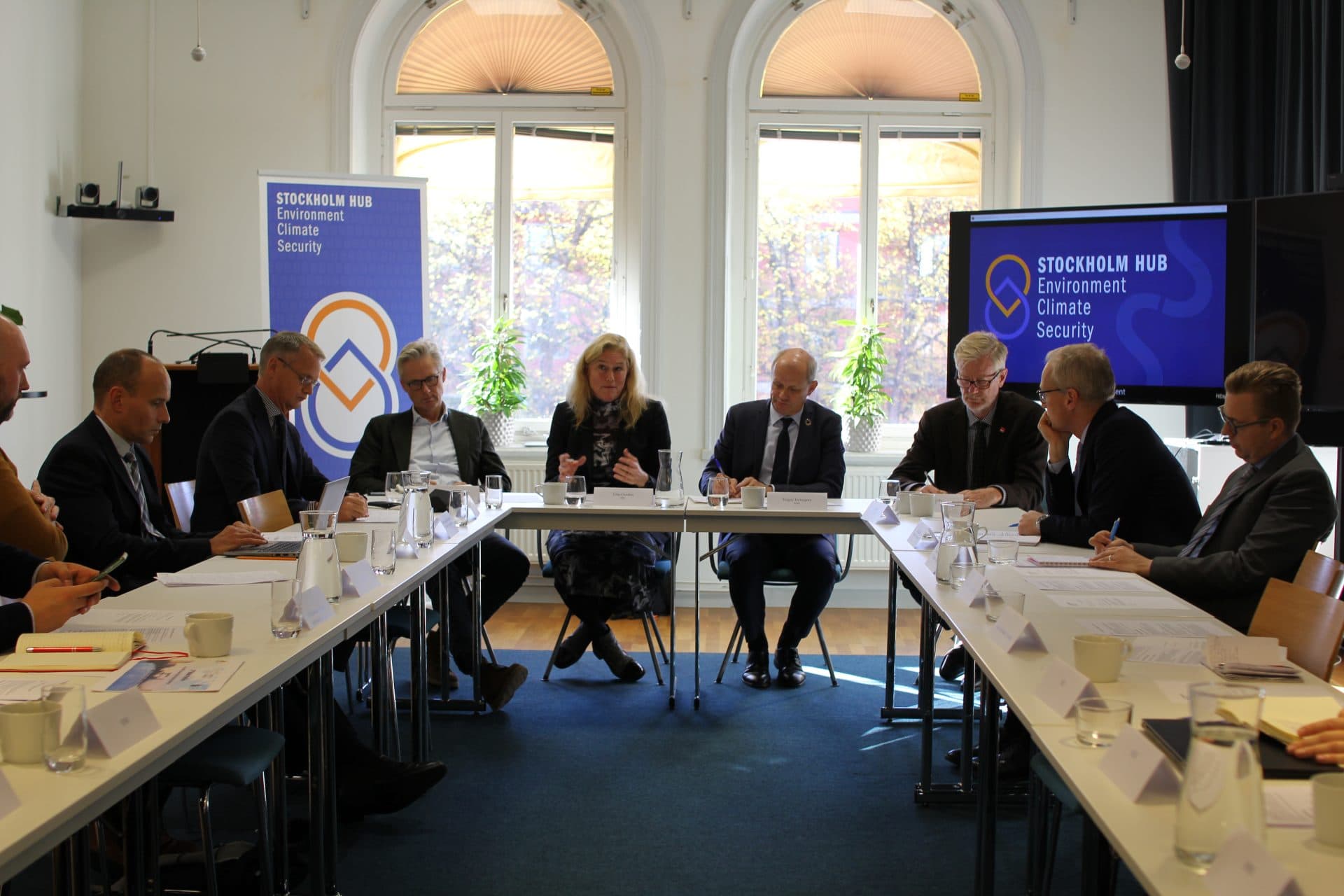 People seated across a table with banners of Stockholm Hub in the background against large white windows. A woman (Line Gordon) is talking and others surrounding (all men) are listening intently.
