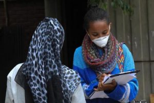 2 women in discussion: one from the back with a head scarf, the other facing her with a face mask, holding a note pad and a pen