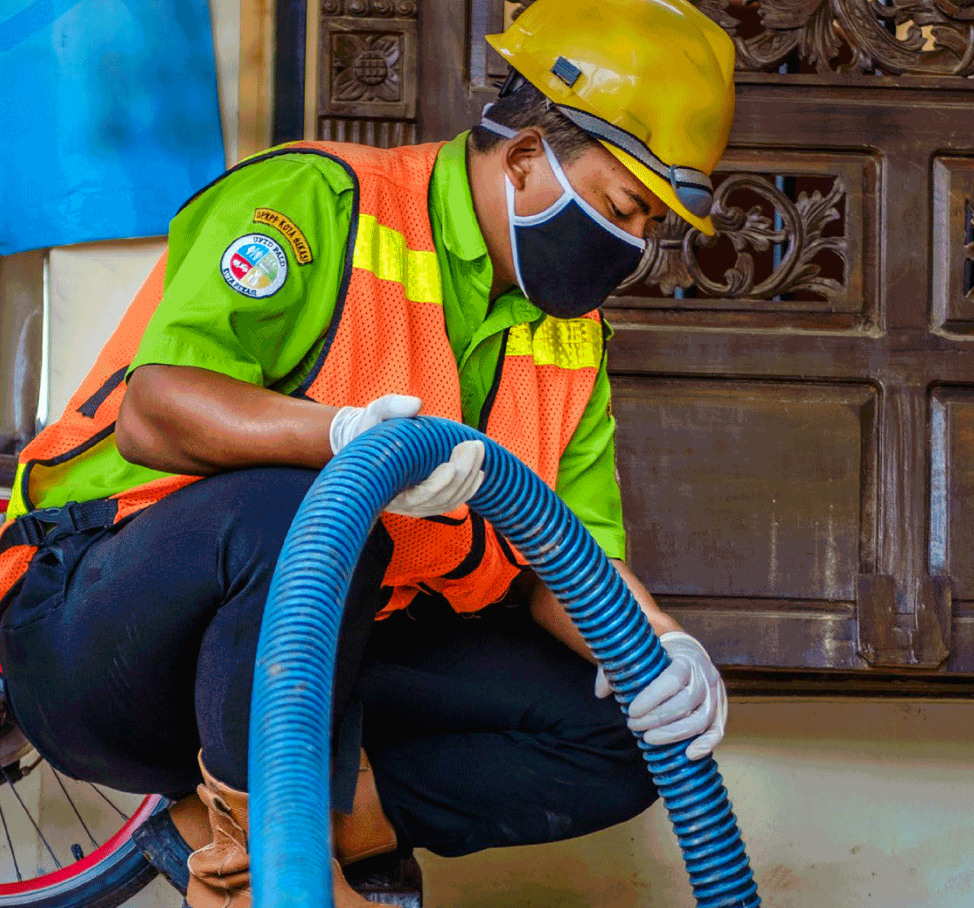 A person wearing a yellow helmet and a black face mask, knee on the floor, holding a blue pipe
