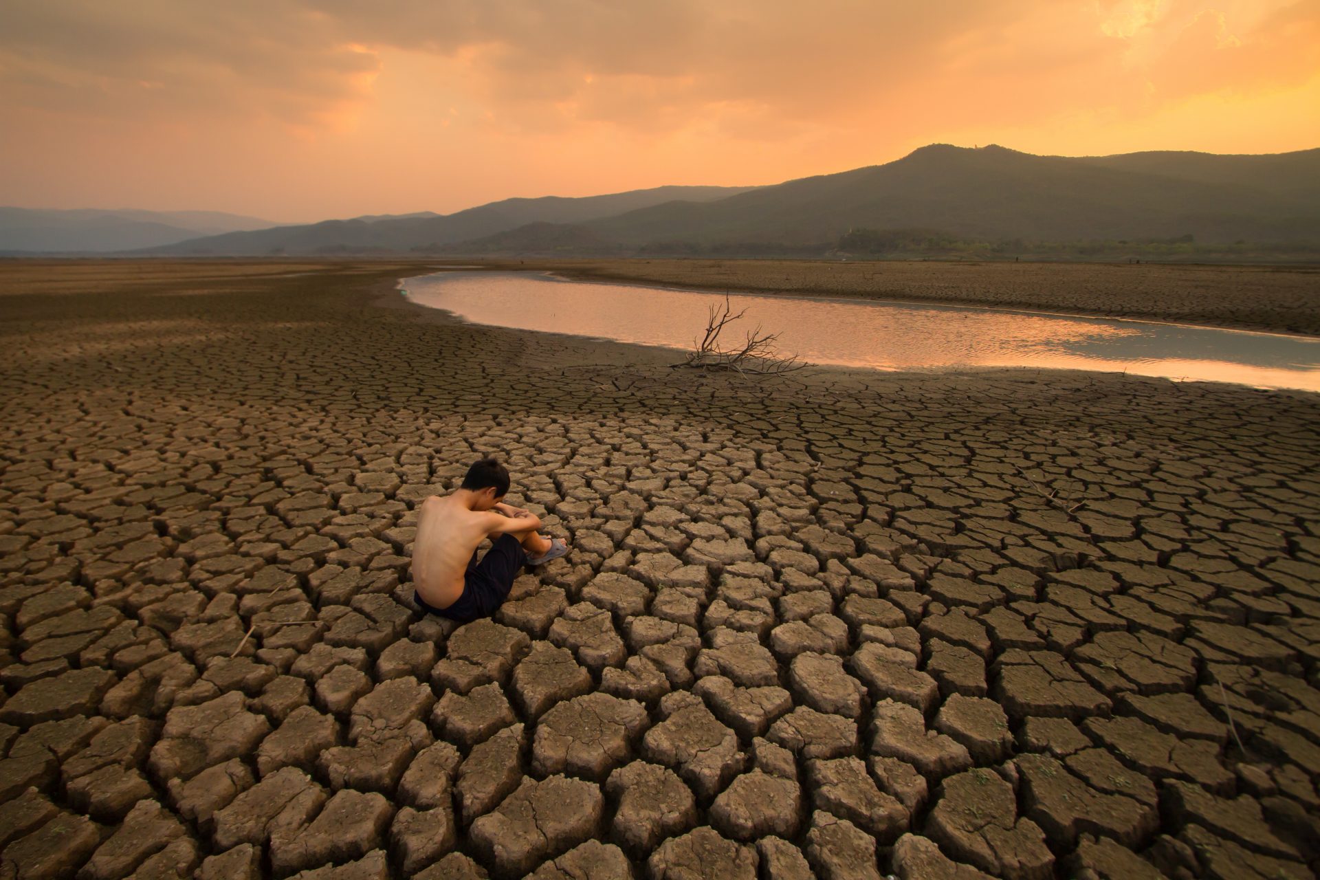 Sad Children or young man sitting on cracked earth near drying river metaphor water crisis, climate change