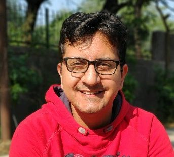 Nitin in a read sweater and glasses against a nature backdrop