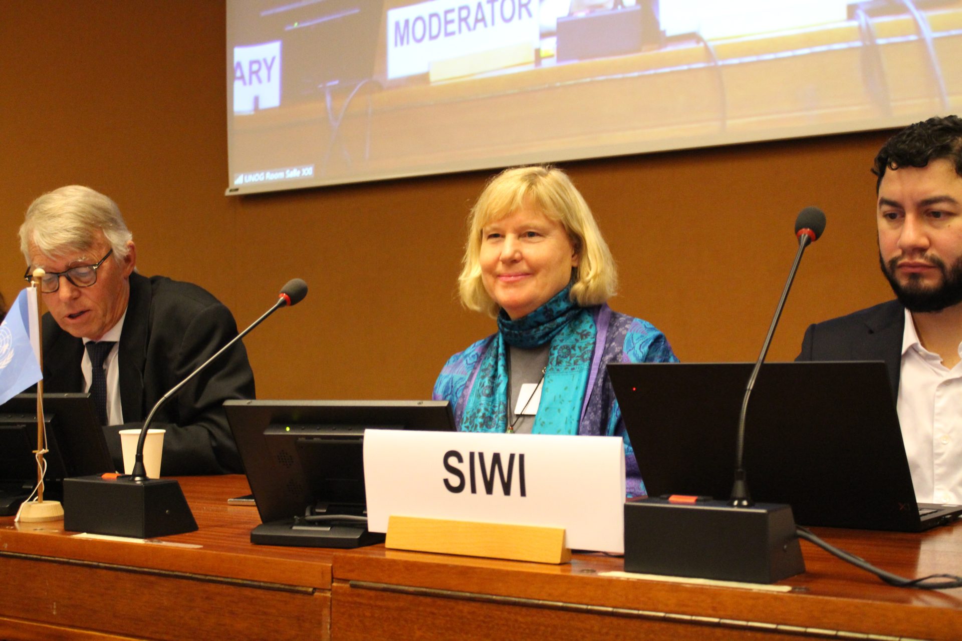 Two people sitting in front of a conference with a screen behind them and sign that states SIWI in front of the table