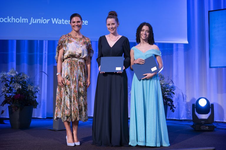 3 women standing up on a stage with blue backlight. On the left is Princess Viktoria in a floral knee length golden dress, in the middle is young Laura in a navy blue floor length gown holding her diploma certificate, and to her right is Camily in a bright blue floor length, off shoulder gown, holding a certificate as well.