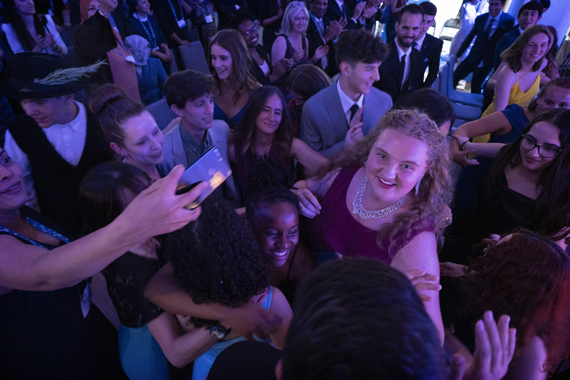 A top view photo of a crowd of young participants. At the centre is a young girl in a red dress and flashy necklace, with wavy golden hair. She is the SJWP winner.