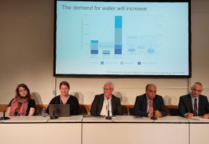 People sitting side by side at a long table in panel, facing camera, at the Bonn Climate Conference 2023. Behind them a large screen with a graph, titled "The demand on water will increase"