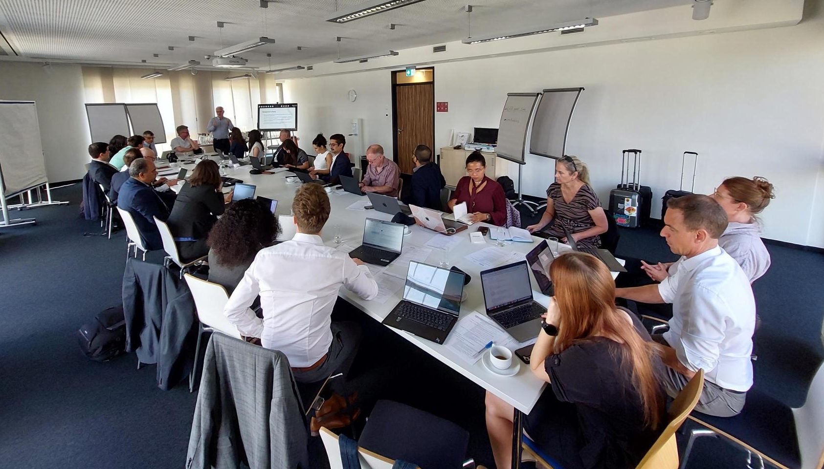 People sitting around a long board table, at the 2023 Bonn Climate Conference. One person standing at the back showing a presentation on the screen