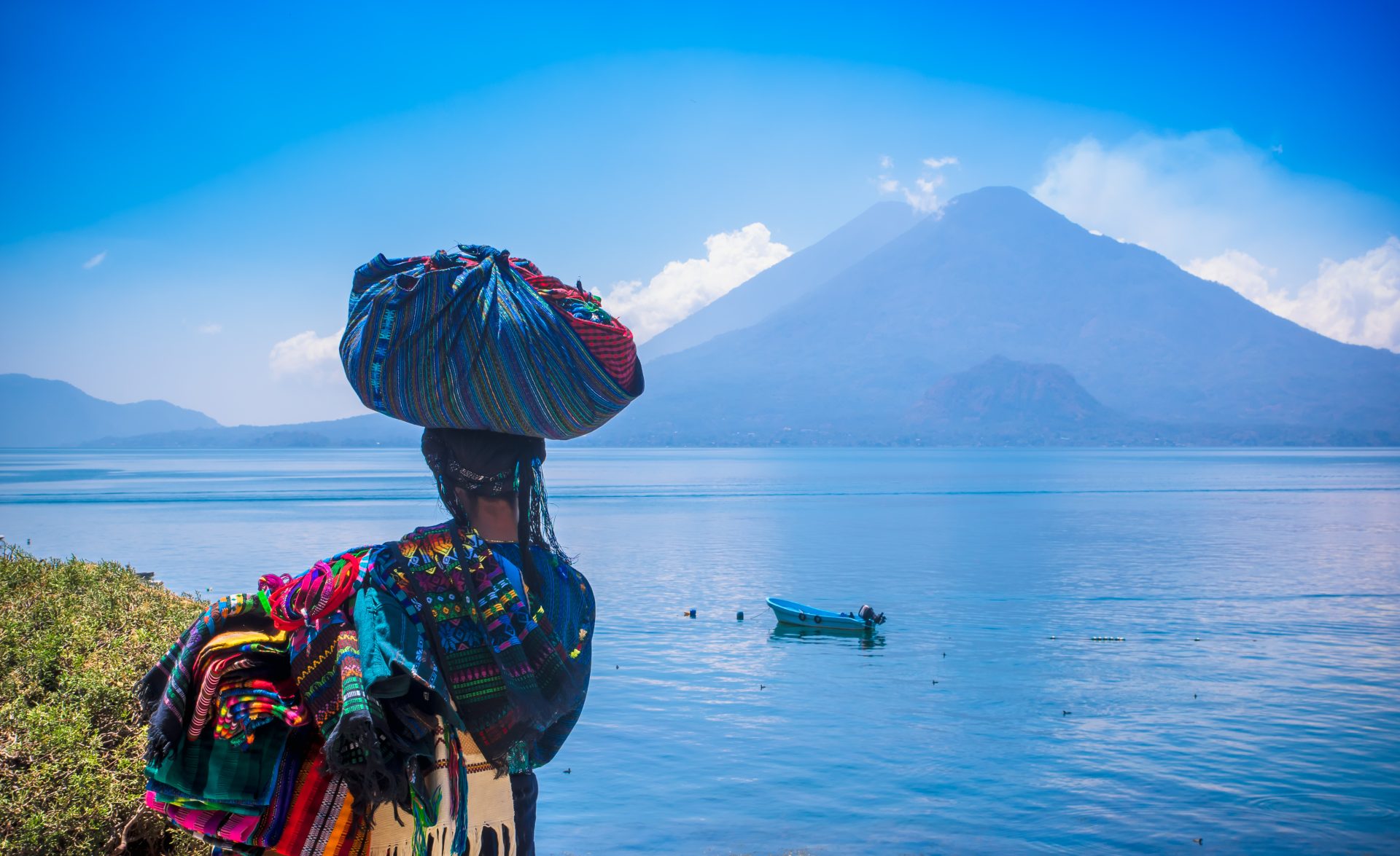 Outdoor view of unidentifed indigenous woman, wearing typical clothes and walking in lakeshore with small boats in Atitlan Lake and volcano in background