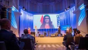 Mishal Faraz winning the People's Choice Award at the Stockholm Junior Water Prize 2022 competition. Ania Anders and Kanika Takar standing on stage, watching a big screen where Mishal's face is projected