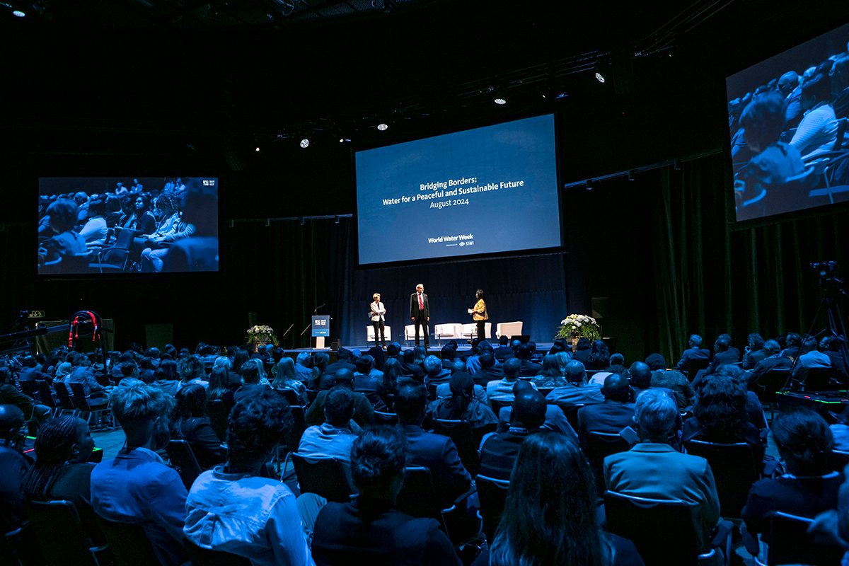 SIWI's Acting Director Karin Gardes, President of World Water Week's Scientific Committee Jon Lane and Host Arati Davis standing on Centre Stage at World Water Week 2023, with large audience sitting in the foreground.